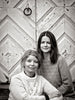 Two women in cozy sweaters sit together before a chevron wooden door. This black-and-white image radiates Scandi holiday warmth, capturing the vibe of Schiffer Publishing, Ltd.'s "Cozy Christmas Crochet.
