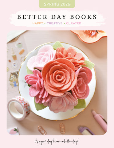 A pastel-themed flat lay featuring a bouquet of pink felt flowers on a plate, surrounded by stationery, washi tape, and paper clips, with the text “Better Day Books: Happy, Creative, Curated. Spring 2026.”.