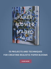 A book titled Paper Flower Magic with a cover showing blue paper flowers and a person in a striped dress, displayed above the text: 15 Projects and Techniques for Creating Realistic Paper Blooms. A Learn More button is below.