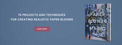 A blue background features text listing “15 projects and techniques for creating realistic paper blooms” next to a book titled Paper Flower Magic with paper flowers on the cover and a pink “Learn More” button below the text.